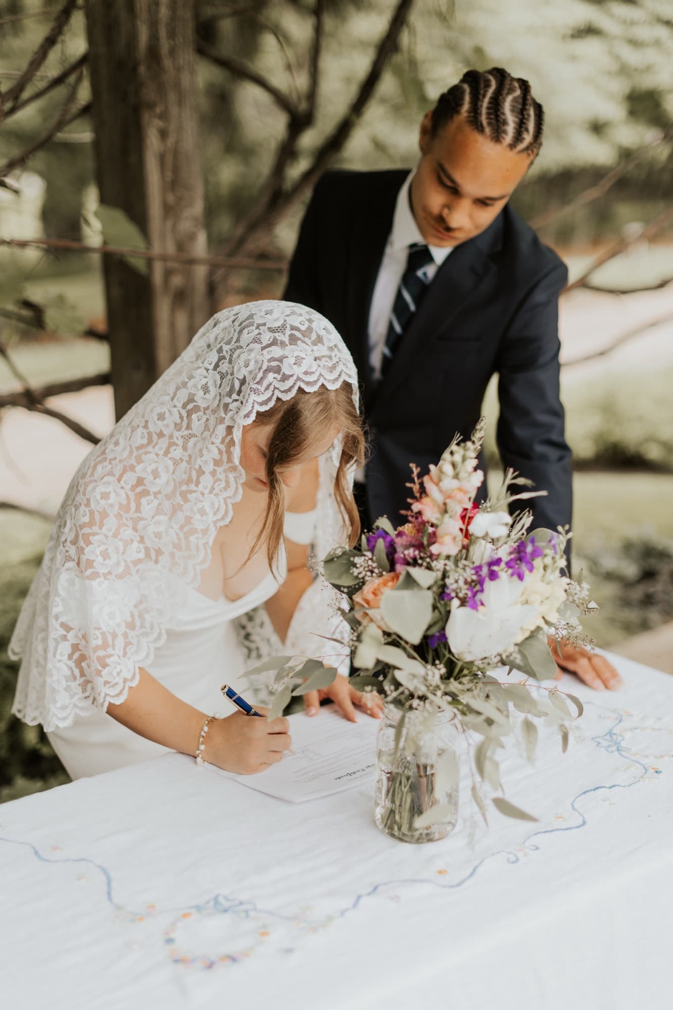 The bride signs the marriage license at a table with a jar of wildflowers as the groom leans in beside her