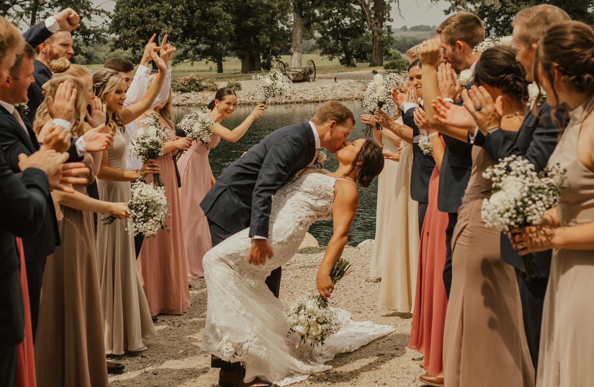A groom dips his bride into a kiss on a gravel path by a pond as the wedding party cheers and raises bouquets on both sides