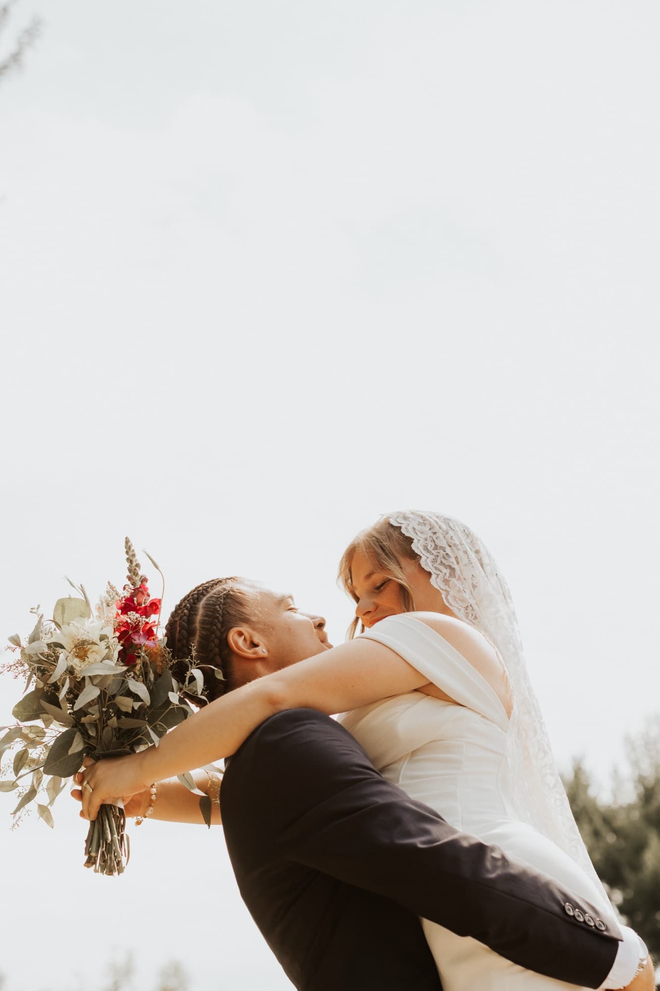 The groom lifts the bride into the air against a pale sky, her wildflower bouquet in her outstretched hand