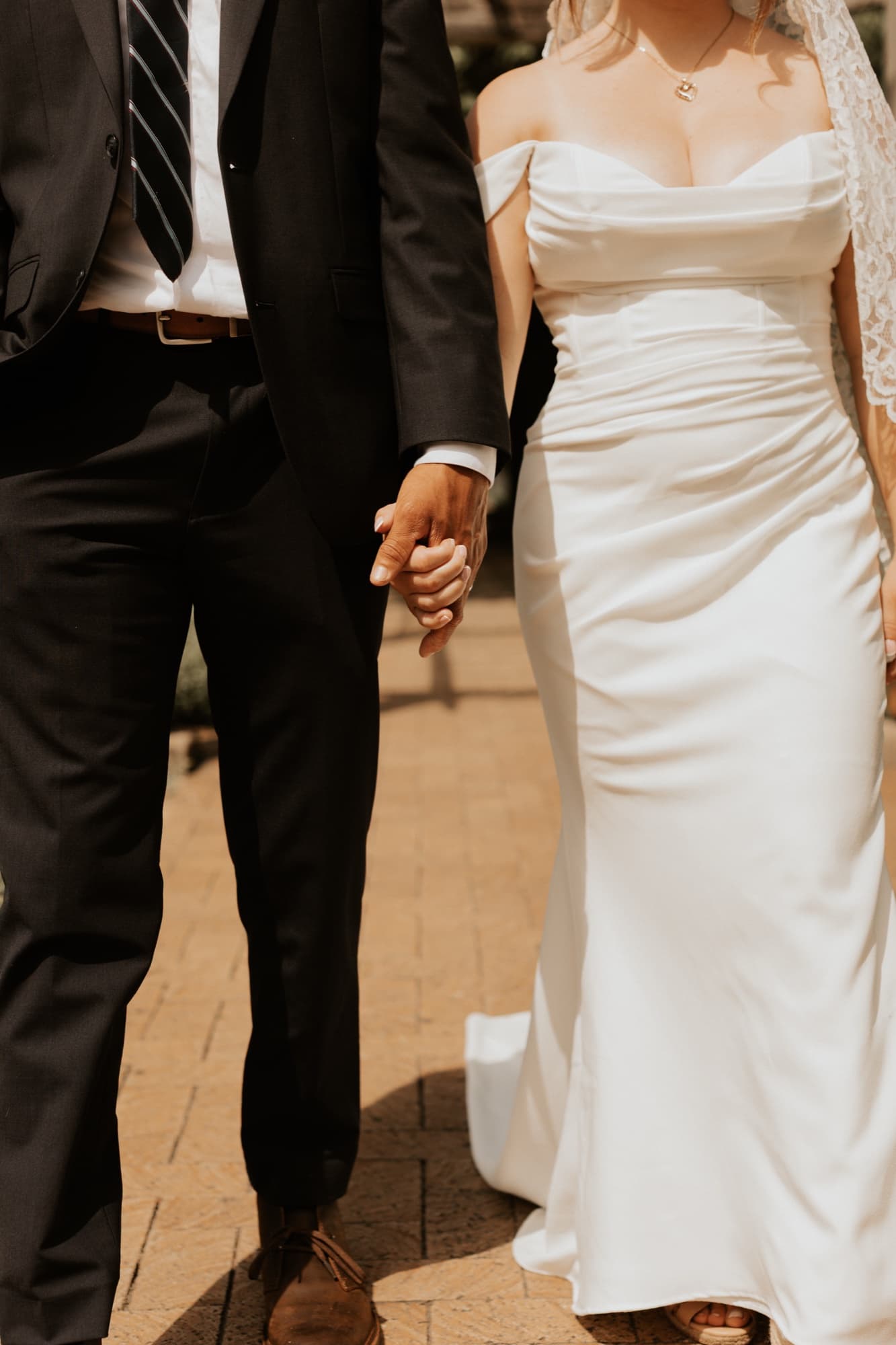 A close crop of Nic and Tirzah walking hand in hand along a brick path, her fitted satin gown catching the light
