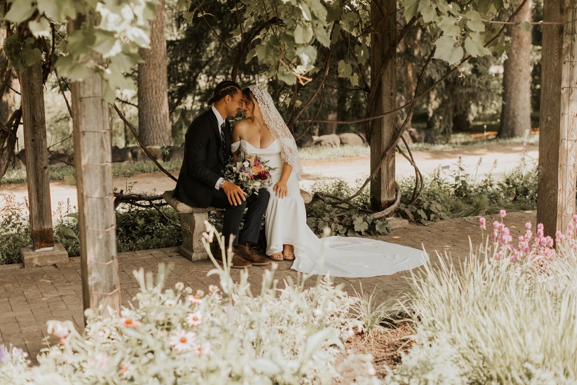 Nic and Tirzah sit close on a garden bench beneath a wooden arbor, surrounded by flowers