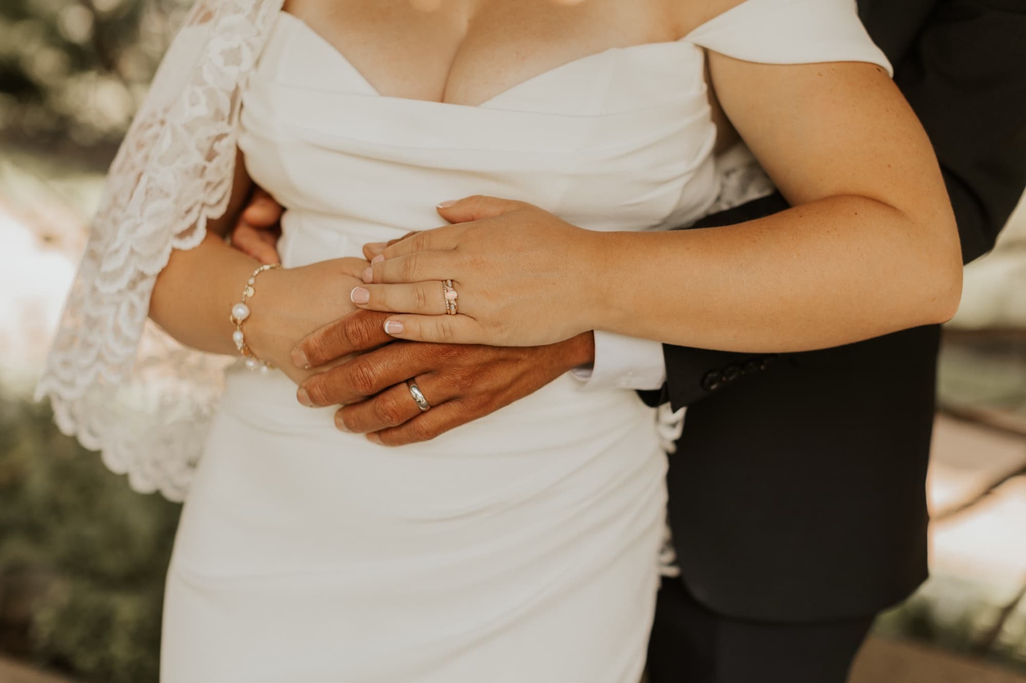The groom's hands rest over the bride's at her waist, both wedding rings showing against her satin gown