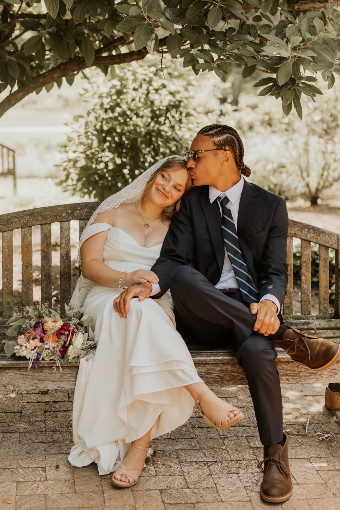 The bride leans her head on the groom's shoulder as he kisses her forehead on a wooden garden bench