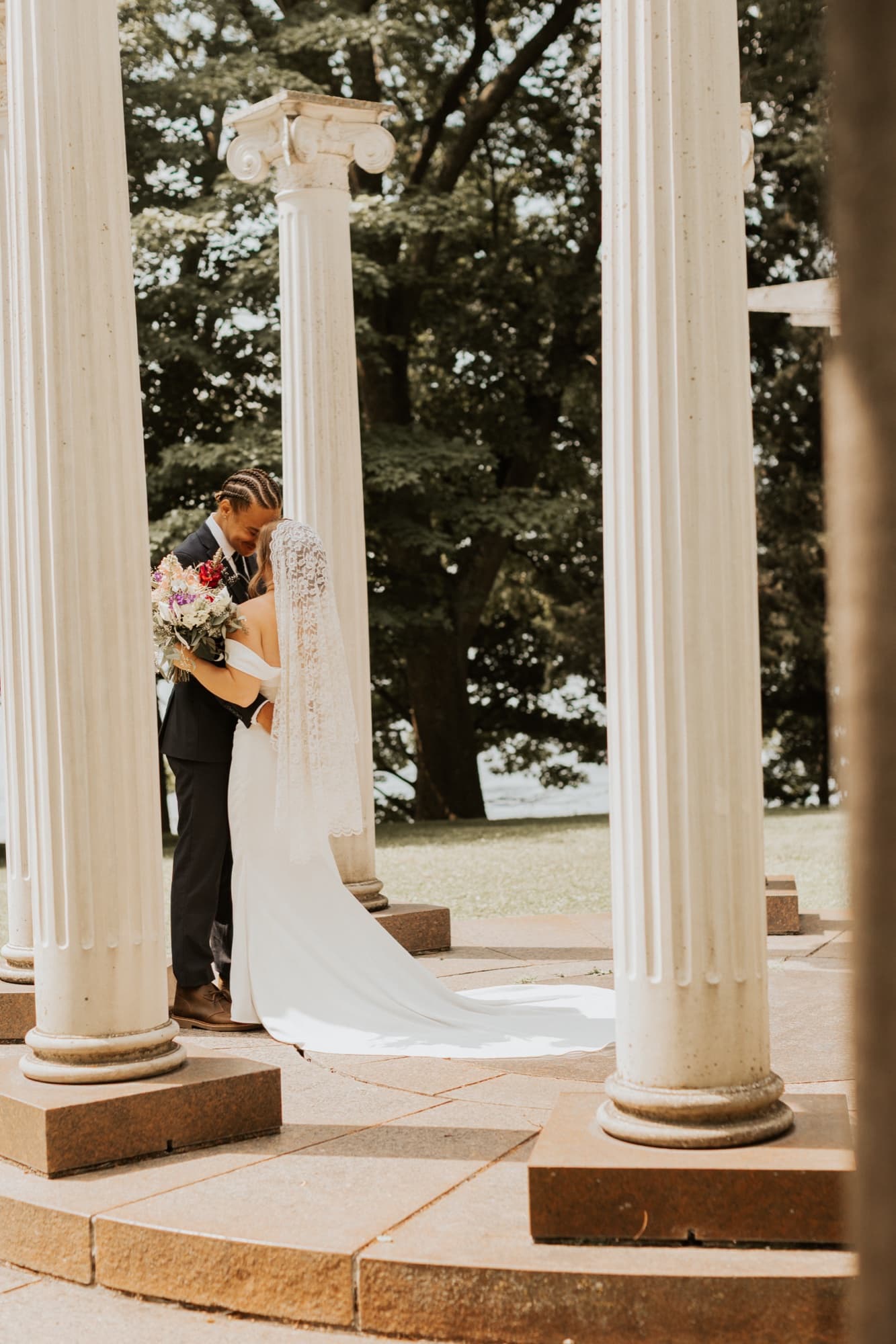 Nic and Tirzah embrace between tall white columns, her veil and train falling across the stone base