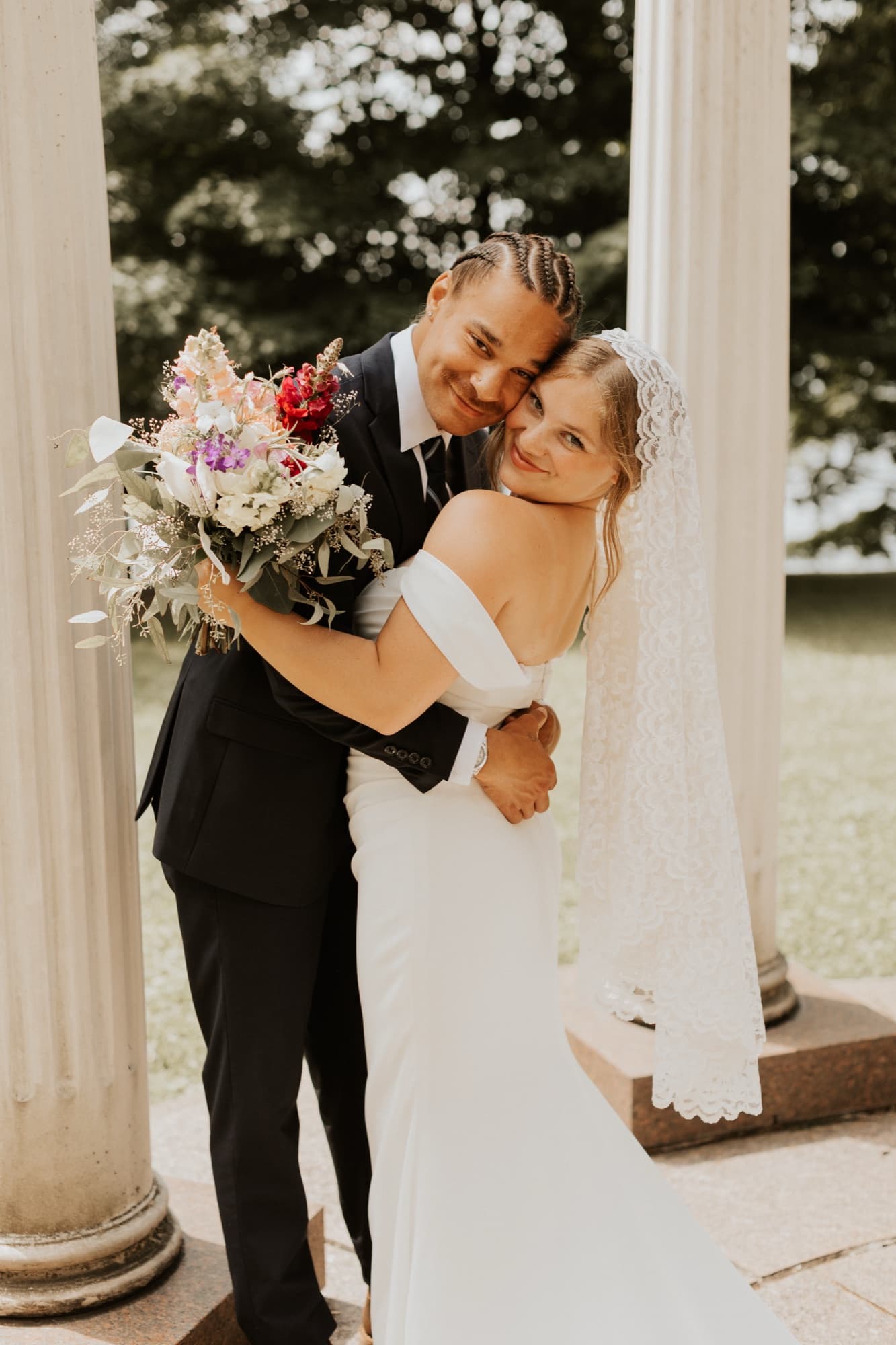 The bride laughs toward the camera as the groom hugs her beside a white column, her wildflower bouquet raised