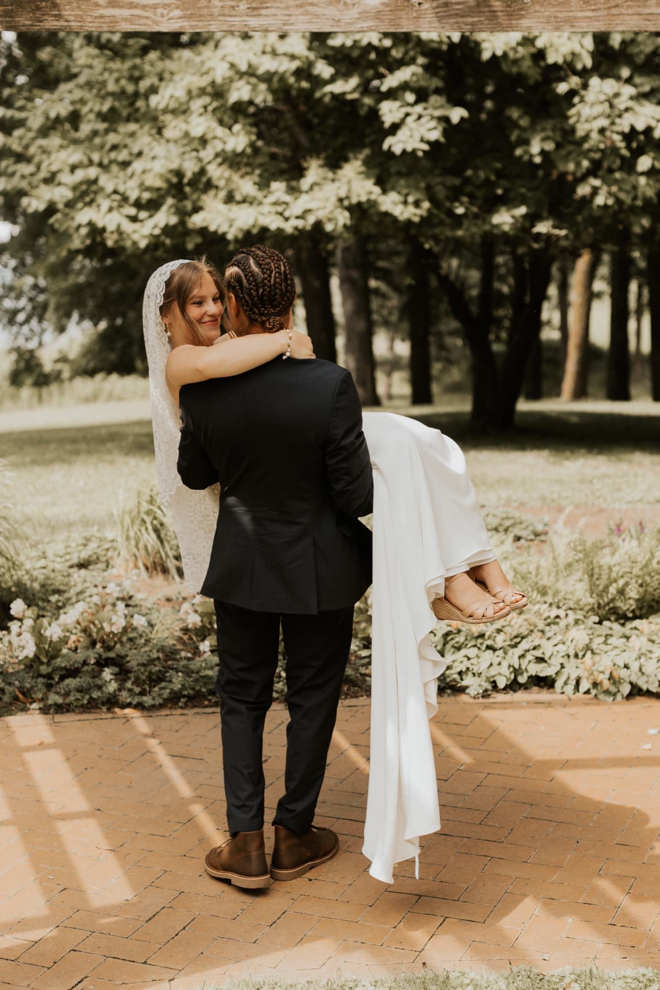 The groom carries the bride away down a brick path, her train hanging over his arm