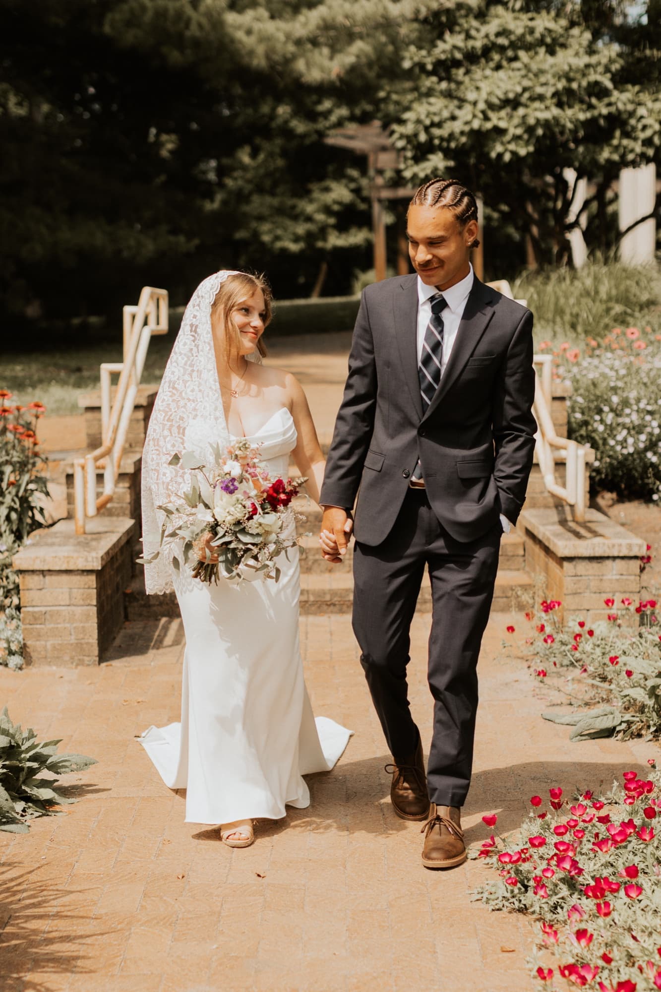 Nic and Tirzah walk hand in hand down a brick path lined with red flowers, smiling at each other