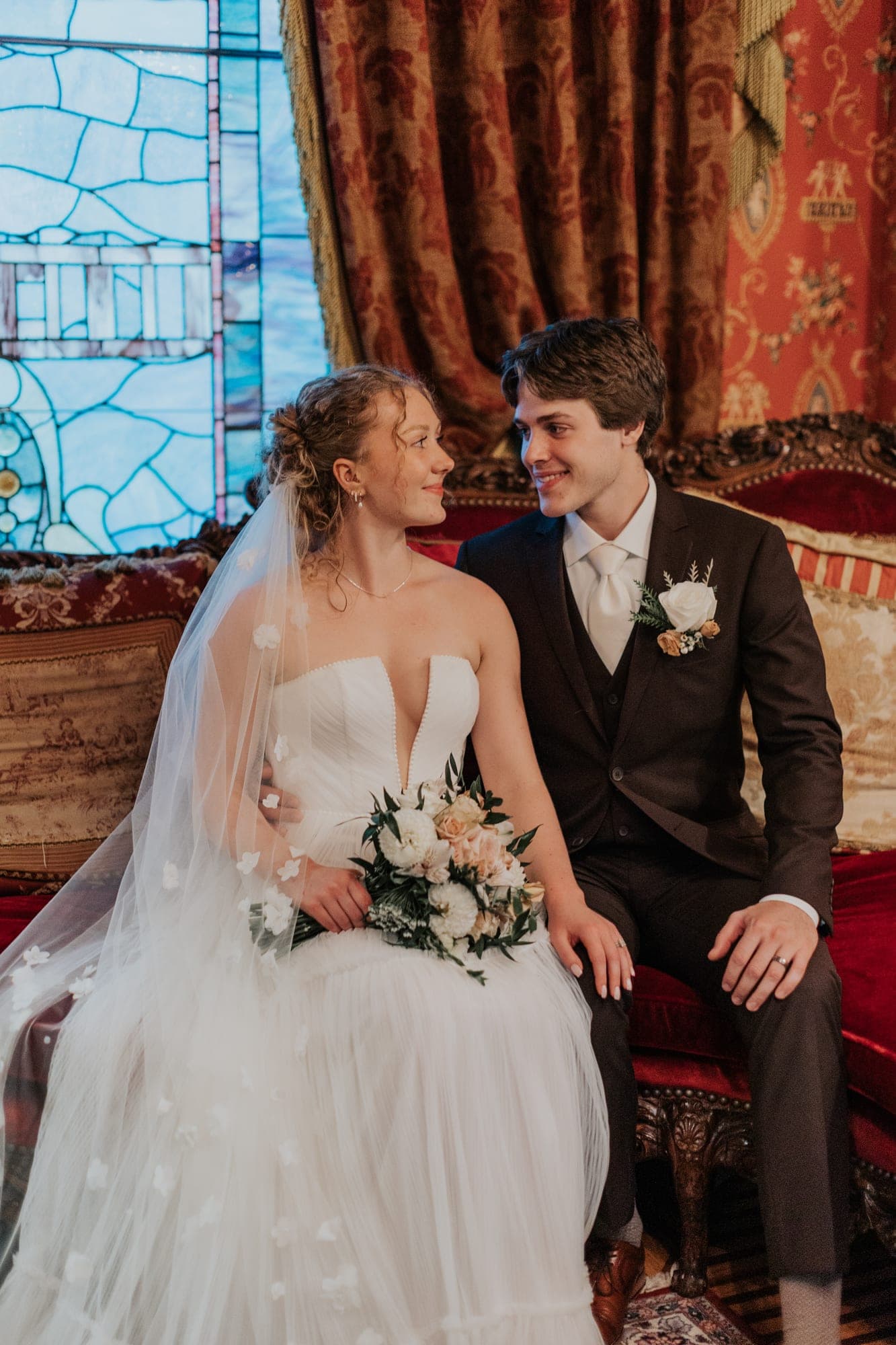 A bride and groom sit close on a red velvet sofa beside a stained-glass window, she holding a white and blush bouquet
