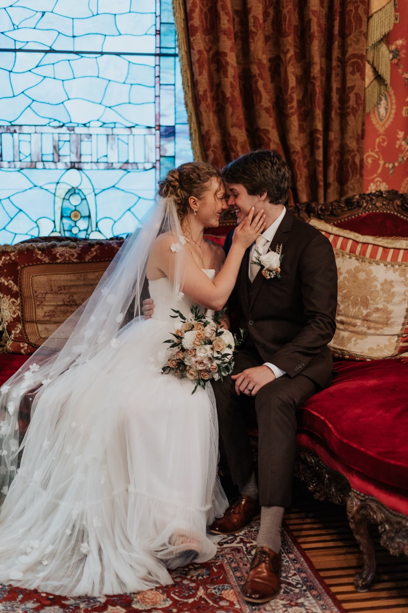 A bride and groom sit close in an ornate room with stained-glass windows and rich red and gold décor