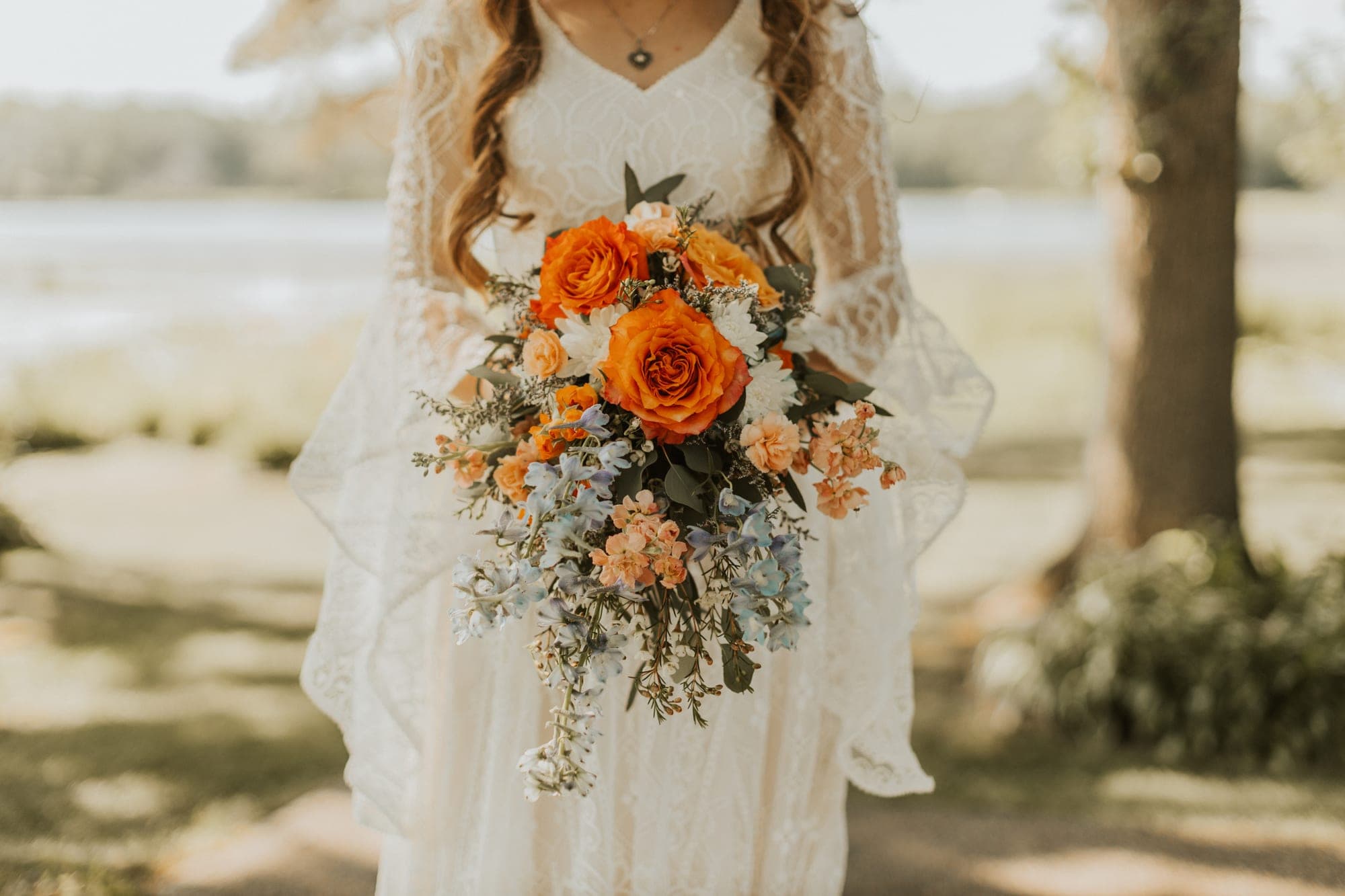 A bride in a lace dress holds a cascading bouquet of orange roses, peach blooms, and blue delphinium in bright backlit sun by a lake