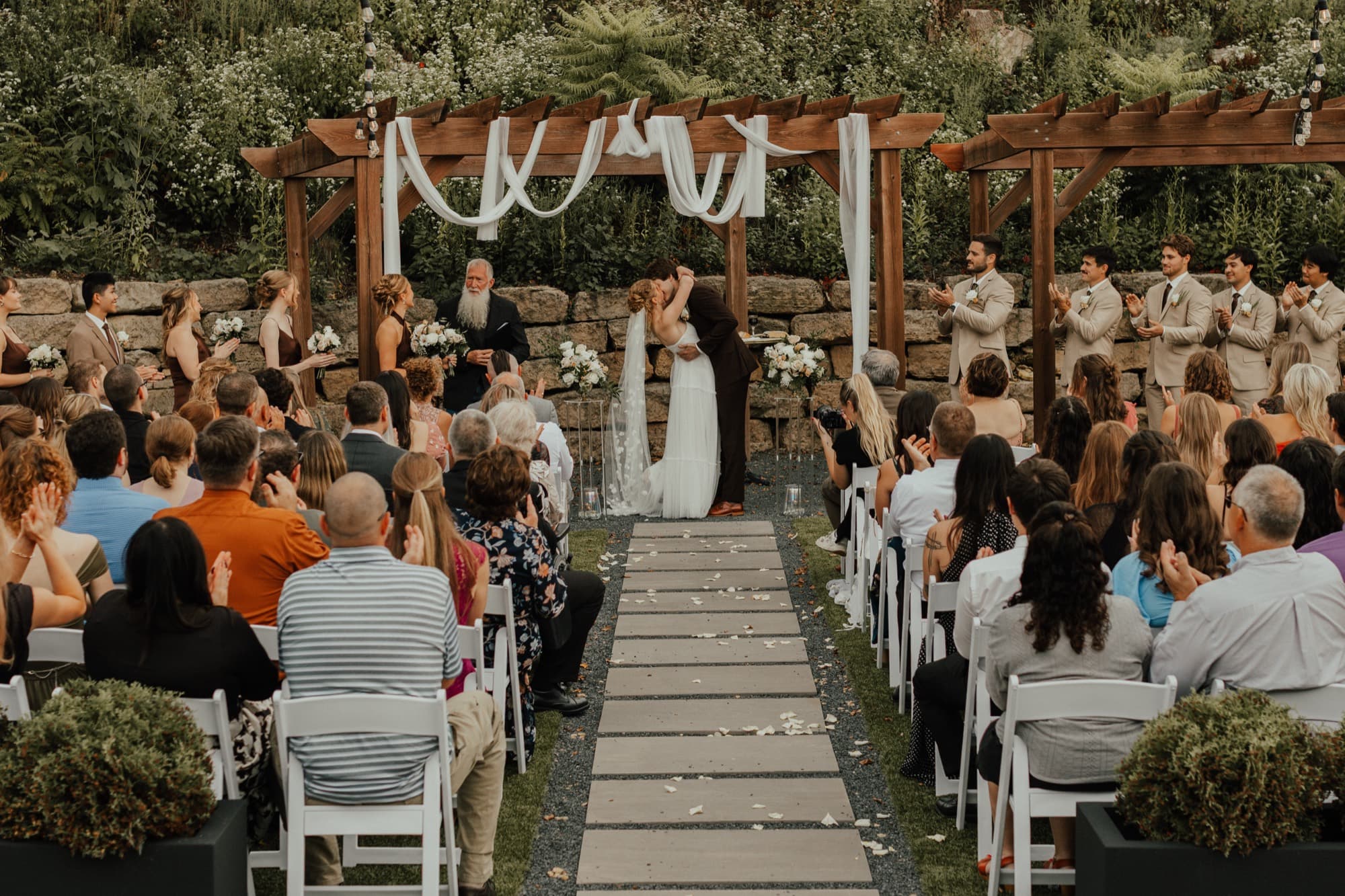 A couple share their first kiss at an outdoor garden ceremony as seated guests applaud