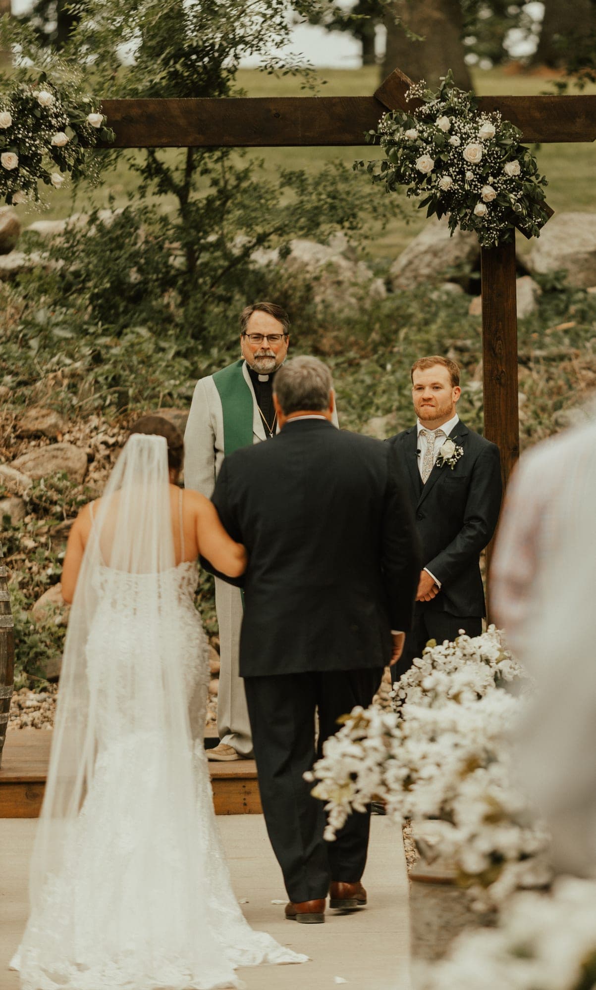 A bride on her father's arm faces the groom and an officiant under a wooden arch dressed with white roses and baby's breath